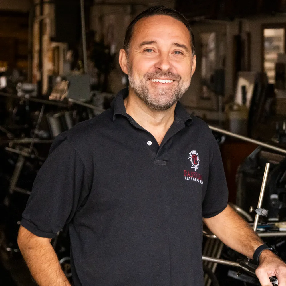 smiling man in a black polo shirt stands in a workshop filled with printing equipment.