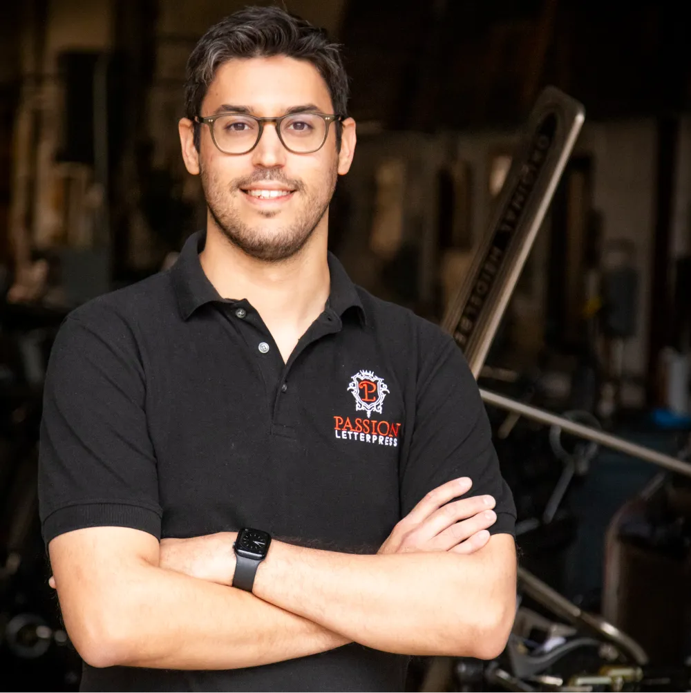 man with glasses stands with arms crossed, wearing a black polo shirt with "Passion Letterpress" logo in a workshop.