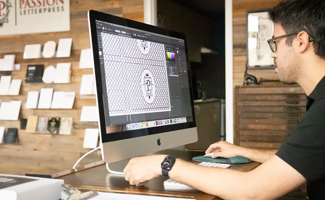 man works on a computer at a desk, designing graphics for a letterpress business in a cozy workspace.