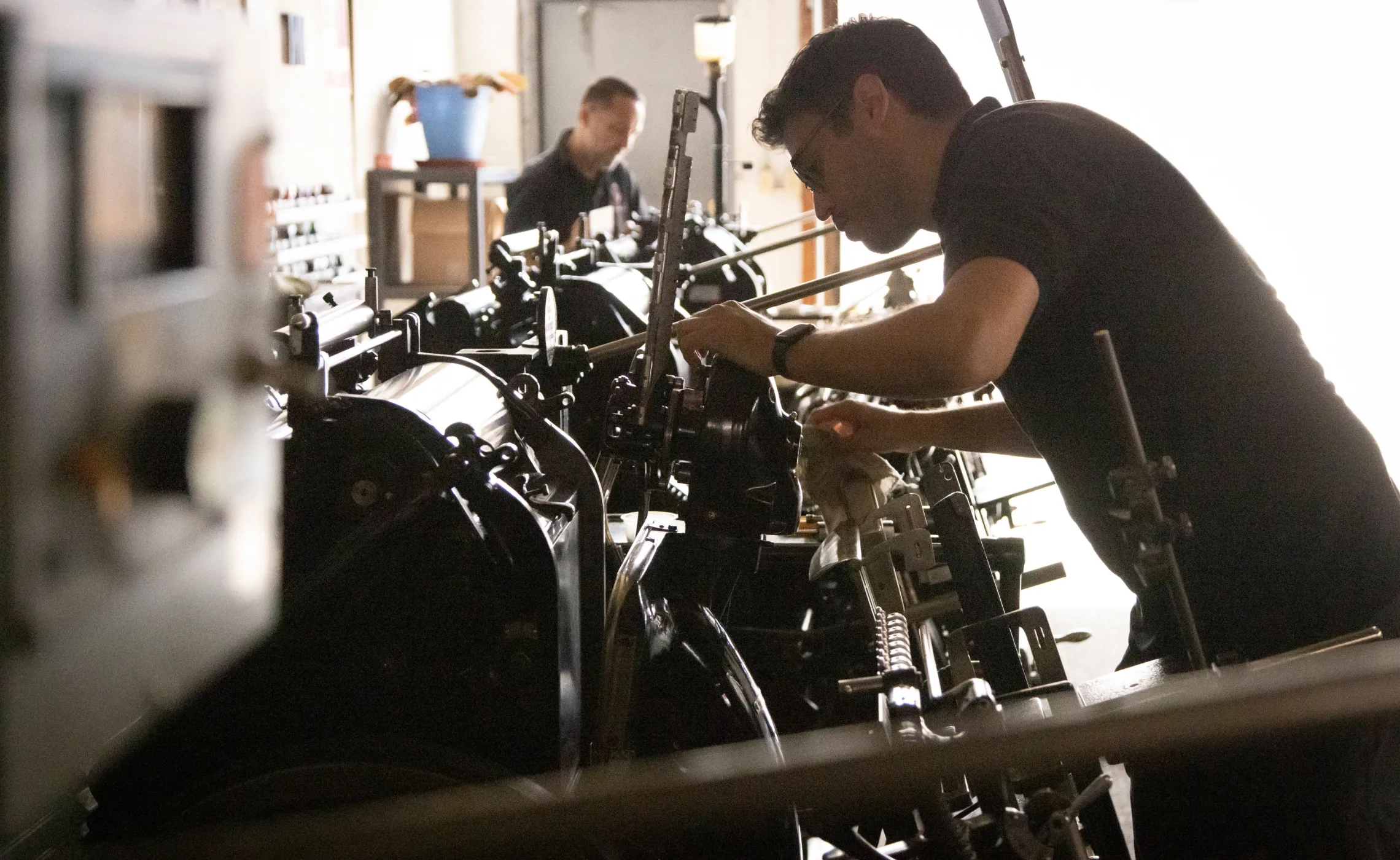 Two men work on machinery in a dimly lit workshop, focusing on mechanical components.