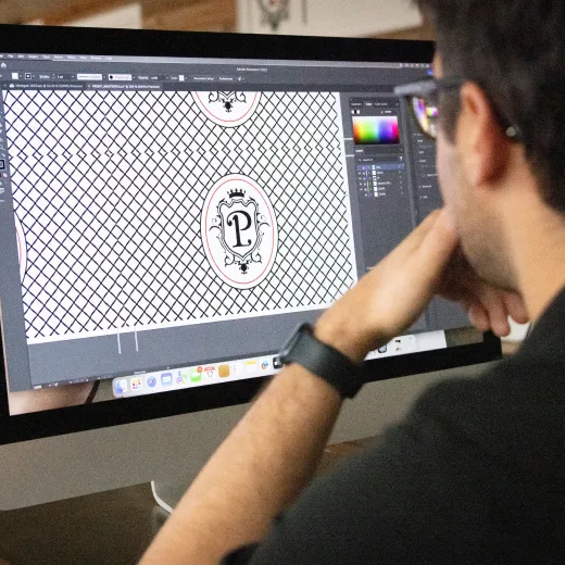 person sits at a computer, focused on graphic design software displaying a patterned design with a logo.